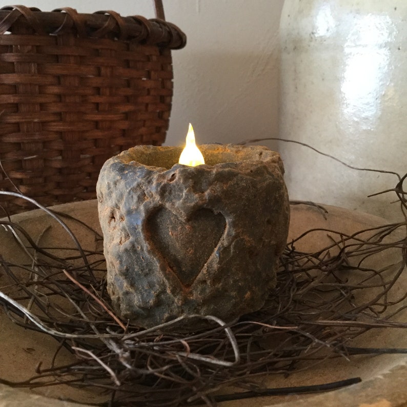 May include: A rustic, battery-operated candle with a carved heart design. The candle has a textured, stone-like appearance and emits a warm, flickering light. It sits in a wooden bowl with twigs, suggesting a natural, home decor theme.