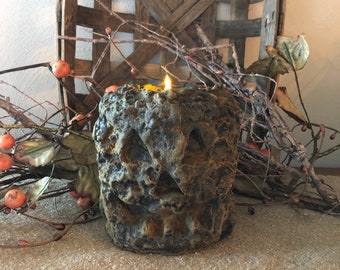Puede incluir: Una vela de color marrón oscuro texturizada con forma de calabaza de Halloween con una vela encendida en su interior.