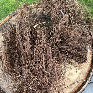 May include: A pile of dried, brown Spanish moss, with a tangled, wiry texture. The moss is resting on a weathered, wooden surface, likely a barrel top. The background shows green grass.