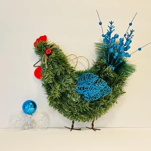 May include: A decorative chicken-shaped wreath made of green faux pine needles, with red comb and wattle accents. It features blue decorative elements and a small blue ornament. The wreath has metal legs and a jute string for hanging.