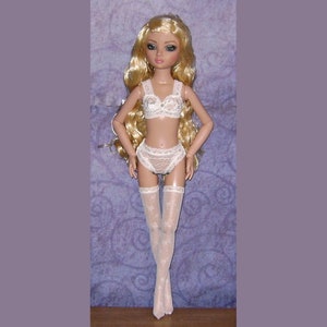May include: A blonde doll wearing a white lace bra and matching panties with white lace stockings. The doll has green eyes and is posed standing with her hands at her sides.