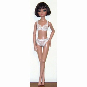 May include: A doll with short black hair and brown eyes is wearing a white lace bra and matching underwear.
