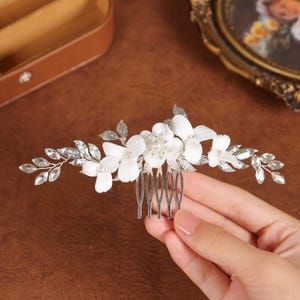 May include: A bridal hair comb with white fabric flowers, faux pearls, and clear crystals. The comb has silver-coloured metal leaves and a row of silver-coloured teeth. The comb is held by a person's hand.