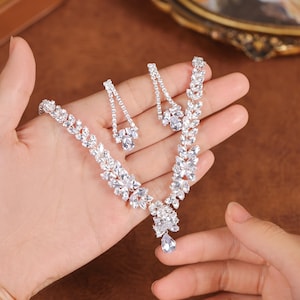 May include: A sparkling jewelry set featuring a necklace and matching earrings. The necklace and earrings are adorned with clear, faceted stones arranged in a floral or leaf-like pattern. The set is displayed on a hand.
