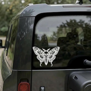 Op de afbeelding: Een witte sticker van een mot met een schedelontwerp op de achterruit van een donkergrijze auto. De mot heeft gedetailleerde vleugelpatronen en een schedel in plaats van de kop. De auto staat buiten met bomen op de achtergrond.
