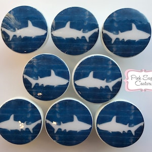 May include: Set of eight white ceramic drawer knobs with a blue and white shark design. The knobs are round and have a distressed woodgrain look.