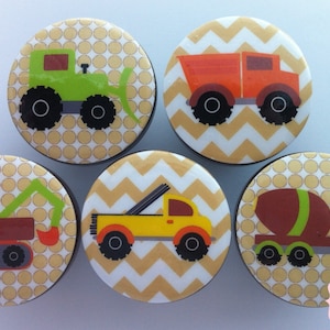 May include: Set of six round knobs with a yellow and white chevron or polka dot pattern. Each knob features a different construction vehicle, including a green bulldozer, orange dumper truck, yellow pick-up truck, brown excavator, brown cement mixer, and a brown dumper truck.