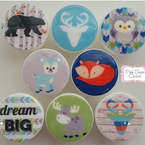Op de afbeelding: Set van acht ronde houten knoppen met verschillende ontwerpen met als thema bosdieren en natuur. De ontwerpen omvatten een hert, een vos, een uil, een beer, een eland en een herten silhouet. Eén knop heeft de tekst "dream big" in een blauw en wit lettertype.