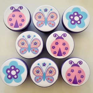 May include: Set of 8 purple and white round knobs with various designs including butterflies, ladybugs, and flowers. The knobs are perfect for adding a touch of whimsy to furniture.