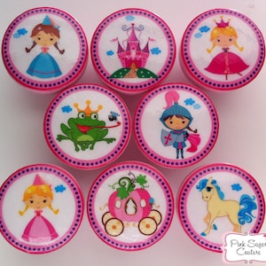 Op de afbeelding: Set van acht ronde roze en witte decoratieve knoppen met verschillende afbeeldingen met een prinsessen-thema, waaronder een prinses, een kasteel, een kikker, een ridder, een pompoenwagen, een paard en wolken.