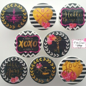 Op de afbeelding: Set van acht ronde zwarte en witte ladeknopjes met gouden accenten. De knoppen hebben verschillende ontwerpen, waaronder een hart met het woord "love", een fiets met bloemen, een sleutel met bloemen, een kroonluchter, een bord met "xoxo", een hart met het woord "love" en de woorden "Hello Gorgeous" en "Ooh la la Paris".