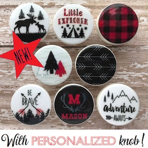 May include: Set of eight round, white ceramic knobs with various woodland-themed designs. The designs include a moose and calf, a tent and trees, a plaid pattern, arrows, a tree, a mountain range, a monogram with antlers, and the words "Be Brave" with arrows. The knobs are new and personalized.