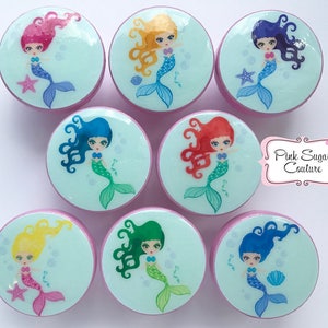May include: Set of eight round, pink-rimmed, decorative knobs with a light blue background and a mermaid design. Each knob features a different colored mermaid with a different colored tail. The mermaids are all smiling and have long hair. The knobs are perfect for adding a touch of whimsy to any room.