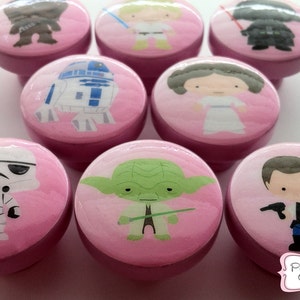 May include: A collection of pink cabinet knobs featuring Star Wars characters like Yoda, Darth Vader, and R2-D2. Each knob has a character illustration on top, with a pink base. The knobs are designed for furniture.