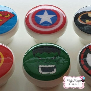 May include: Six white cabinet knobs with superhero designs. The knobs feature Superman, Captain America, Batman, The Flash, The Hulk, and Spider-Man. The knobs are round with a glossy finish, perfect for a child's bedroom or a themed space.