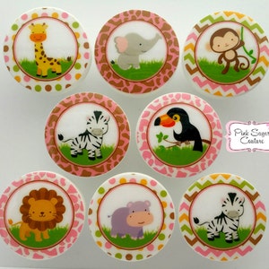 以下が含まれることがあります： ピンク、茶色、緑の動物のイラストが描かれた、丸い白いセラミックノブ8個セット。動物には、キリン、ゾウ、サル、シマウマ、オニオオハシ、ライオン、カバ、シマウマが含まれています。各ノブには、異なる動物と、縁の周りに異なるパターンが描かれています。