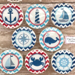 May include: Set of 8 nautical themed ceramic round ornaments with red and blue chevron patterns. Each ornament features a different nautical icon: anchor, lighthouse, sailboat, compass, whale, crab, ship's wheel, and another sailboat.