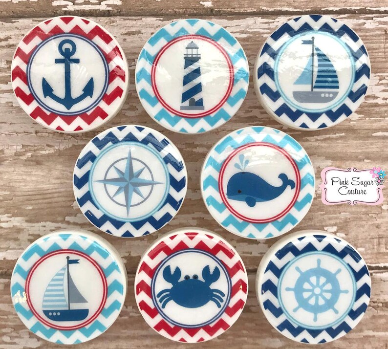 May include: Set of 8 nautical themed ceramic round ornaments with red and blue chevron patterns. Each ornament features a different nautical icon: anchor, lighthouse, sailboat, compass, whale, crab, ship's wheel, and another sailboat.