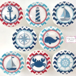 Puede incluir: Conjunto de ocho pomos redondos de cerámica con temática náutica con patrones de chevron rojo y azul. Cada pomo presenta una imagen náutica diferente: ancla, faro, velero, brújula, ballena, cangrejo, timón y velero.