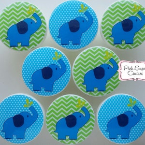 Könnte beinhalten: Satz mit 8 runden Schubladengriffen im Elefanten-Design in Blau und Grün. Jeder Griff zeigt einen blauen Elefanten mit einem grünen Blatt im Rüssel vor einem Hintergrund aus blauen Punkten oder grünem Zickzackmuster.