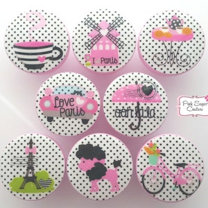 Peut inclure: Ensemble de huit boutons en polka dots rose et blanc avec différents motifs sur le thème parisien, dont un moulin à vent, la tour Eiffel, une voiture qui dit "Love Paris", un caniche, un vélo, un service à thé, un parapluie et une tasse de café.