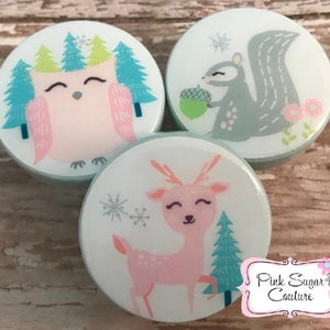 Puede incluir: Tres pomos redondos de cerámica blanca con un borde azul claro. Cada pomo presenta un animal de bosque diferente: un búho, una ardilla y un ciervo. El búho es rosa con los ojos cerrados, la ardilla es gris con una bellota verde y el ciervo es rosa con manchas marrones. Los pomos están decorados con copos de nieve y pinos.