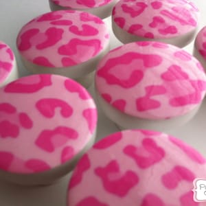 Puede incluir: Primer plano de varios dulces rosas y blancos. Las tapas están decoradas con un estampado de leopardo rosa brillante. Los dulces son redondos y se asientan sobre una base blanca. La imagen está bien iluminada y los dulces están enfocados.