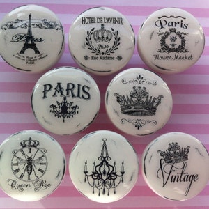 May include: Eight white ceramic knobs with black and white designs. The designs include a crown, a bee, a chandelier, and the word "Paris".