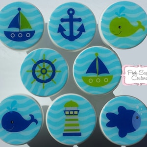 May include: Set of nine nautical themed cabinet knobs with blue, green, and white colors. The knobs feature images of a sailboat, anchor, wheel, whale, lighthouse, and fish.