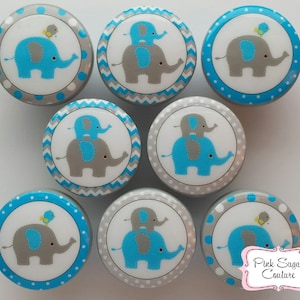 Könnte beinhalten: Acht runde blaue und graue Knöpfe mit weißem Hintergrund und einem Design eines blauen Elefanten mit einem kleineren grauen Elefanten darauf. Die Knöpfe haben ein graues, weißes und blaues Zickzackmuster oder ein graues, weißes und blaues Punkte-Muster am Rand.