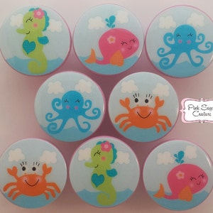 Könnte beinhalten: Satz von acht bunten Keramik-Knöpfen mit Meeresbewohnern. Die Knöpfe sind rund und haben einen hellblauen Hintergrund mit weißen Wolken. Die Lebewesen umfassen ein Seepferdchen, einen Wal, einen Oktopus und eine Krabbe. Jeder Knopf hat ein süßes, cartoonartiges Gesicht.