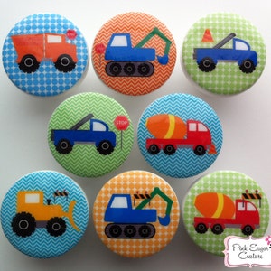 May include: Set of eight colourful round knobs with different construction vehicle designs. Each knob features a different vehicle, including a dumper truck, cement mixer, excavator, and tow truck. The knobs have a blue, green, orange, and red background with white polka dots or chevron patterns.
