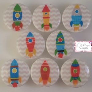 May include: Eight round, white, decorative buttons with a chevron pattern. Each button features a colorful rocket ship with a different color scheme. The text "Pink Sugar Couture" is visible in the background.