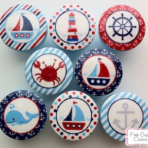 Op de afbeelding: Negen ronde, blauw-witte nautische knoppen met verschillende ontwerpen, waaronder een vuurtoren, een zeilboot, een anker, een krab, een walvis en een stuurwiel. De knoppen zijn versierd met rode, blauwe en witte strepen, stippen en ankers.