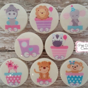 Könnte beinhalten: Satz von acht runden, weißen, keramischen Cake Pops, die mit verschiedenen Tieren auf einem Zug verziert sind. Zu den Tieren gehören ein Löwe, ein Zebra, ein Elefant, eine Giraffe, ein Affe, ein Tiger, ein Nilpferd und ein Zug. Die Cake Pops sind mit Rosa, Lila, Blau und Weiß verziert.