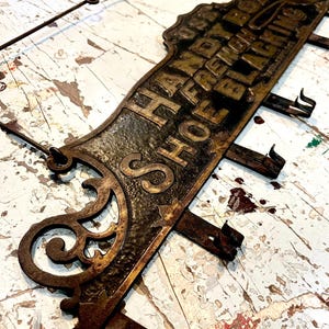 Puede incluir: Un letrero antiguo de hierro fundido con las palabras "HANDY BOY SHOE BLACKING" en letras doradas en relieve. El letrero tiene un diseño ornamental y está muy desgastado con óxido.