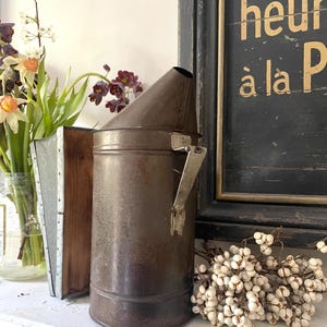 May include: A rustic metal bee smoker, featuring a conical top and a handle, in a weathered brown finish. It rests on a white surface, with a wooden box of flowers and a framed sign visible in the background.