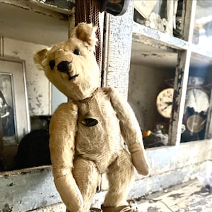 Puede incluir: Un oso de peluche de estilo vintage, de color marrón claro, con una campana alrededor del cuello. El oso tiene ojos negros y una nariz negra. Presenta un aspecto desgastado y antiguo. El fondo incluye una foto enmarcada y un reloj.