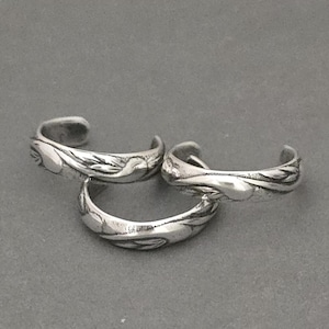 May include: Three silver toe rings with a floral design. The rings are stacked on top of each other.