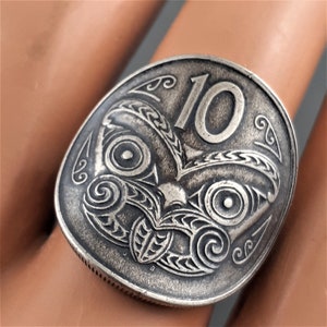 May include: A silver coin ring featuring a Maori design with a stylized face and the number 10.