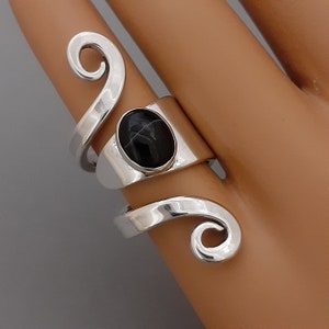 May include: A silver ring with a black oval stone set in the center. The ring is designed with two swirling bands that wrap around the finger.