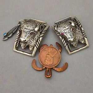 May include: Two silver belt buckles with a buffalo head design. The buffalo has red eyes and a textured fur. One buckle has a turquoise feather charm.