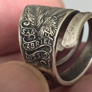 May include: A silver-toned ring with the words "SAN GABRIEL MISSION" engraved on a banner. The ring features a detailed palm tree design. The ring is wrapped in a spiral design.