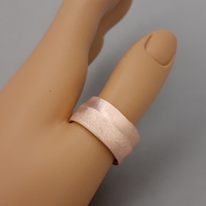 May include: A rose gold colored ring with a smooth, slightly textured surface. The ring is worn on a finger.