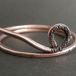 May include: A copper bangle bracelet with a textured, curved design. The bracelet is made of two pieces of copper wire that are intertwined.