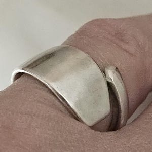 May include: A silver ring with a wide, curved band and a smaller, curved accent. The ring has a smooth, polished surface and a modern design. The ring is worn on a finger.