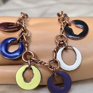 May include: A copper-toned bracelet featuring a series of colorful, enamel-coated circular charms. The charms are in shades of blue, yellow, purple, white, black, and red. The bracelet is composed of linked rings and twisted copper wire.