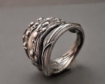 May include: A silver ring with a floral design. The ring appears to be crafted from a spoon, with the spoon's handle forming the band. The floral design features small, round buds and delicate leaves.