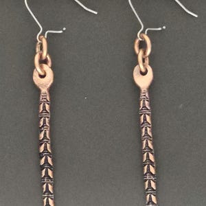 May include: A pair of copper-colored earrings with a key-like design. Each earring features a decorative, elongated pendant with a series of repeating geometric shapes. The earrings hang from a simple, curved ear wire.