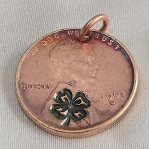 May include: A copper penny with a four-leaf clover design embedded in the center. The penny is dated 1953 and has the words "In God We Trust" and "Liberty" inscribed around the edge. The penny has a loop attached to the top for hanging.
